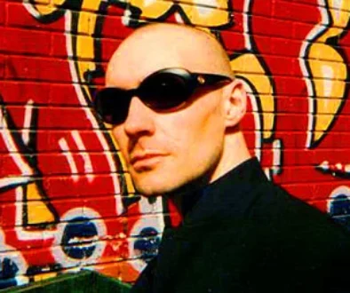 Grant Morrison, bald and in sunglasses, in front of a graffiti mural