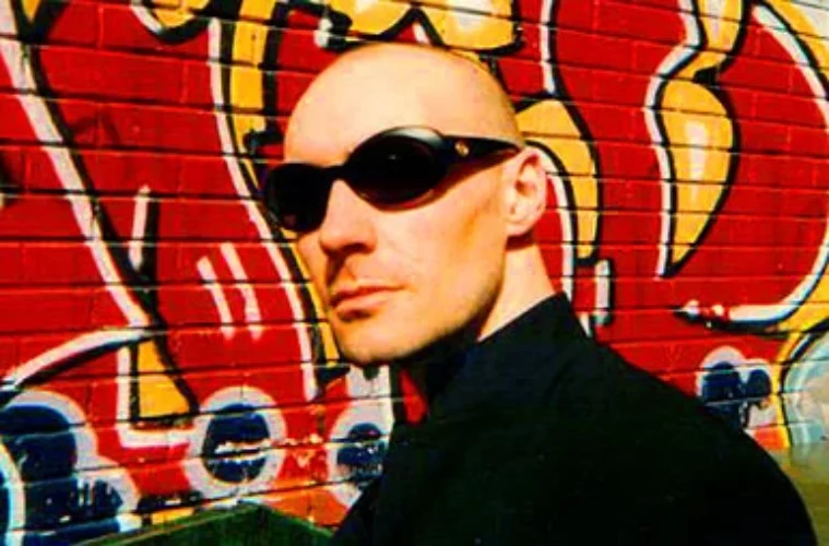 Grant Morrison, bald and in sunglasses, in front of a graffiti mural