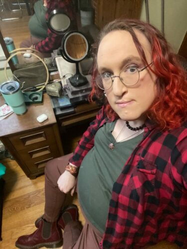 A middle-aged trans woman with dyed red hair is wearing an olive green t-shirt, red and black flannel, brown denim pants, and red Doc Martens Chelesa boots. Her necklace is a copper apple pendant, and she is wearing a copper bracelet and earrings. Her other necklace is made of blue stone.